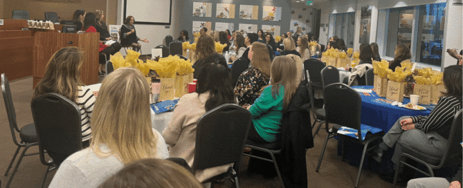 A panel speaker addresses a room of seated women at a professional event with decorated tables.