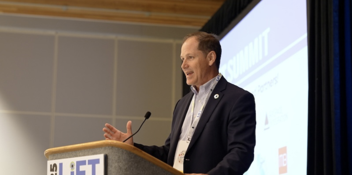 Josh Metz speaking at a podium during a summit, with a presentation screen and event logos in the background.