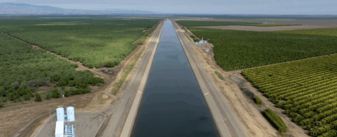 A wide canal running straight through farmland, with green orchards and crop fields on both sides under a cloudy sky.