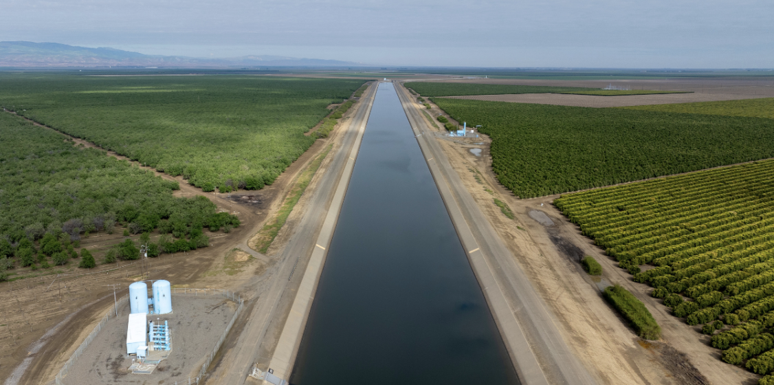 A wide canal running straight through farmland, with green orchards and crop fields on both sides under a cloudy sky.