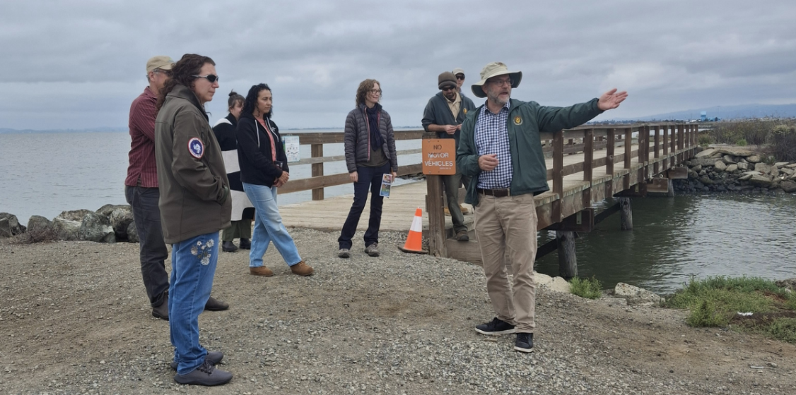 A group of people standing near a pier by the shoreline, listening to a man in a hat and jacket who is speaking and gesturing toward the water.