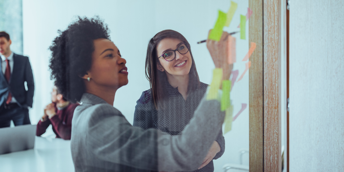 Two professionals collaborate at a glass wall, reviewing and writing notes on colorful sticky notes during a team planning session.